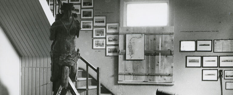 Puinen laivan keulakuva ja kehystettyjä kuvia esillä näyttelytilassa.