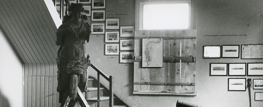 Puinen laivan keulakuva ja kehystettyjä kuvia esillä näyttelytilassa.